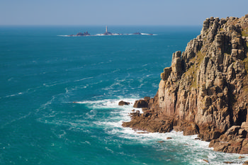 Longships lighthouse cliffs close This is a landscape photograph capturing the rugged cliffs near Longships Lighthouse along the coast of Cornwall, United Kingdom. Taken during the afternoon in early spring, the image shows the striking natural rock formations as they rise sharply from the turquoise waters of the Atlantic Ocean. Longships Lighthouse, an important maritime landmark, is visible in the distance standing on rocky islets. The scene highlights the dramatic nature of the Cornish coastline, with the powerful waves breaking against the base of the cliffs, illustrating the untamed beauty of nature in this region of the United Kingdom.